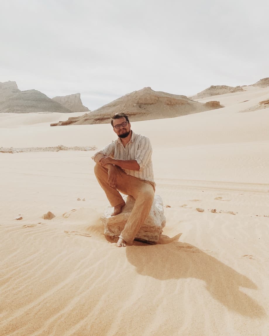 A person seated on a rock in a quiet desert landscape, with sand dunes and distant rock formations.